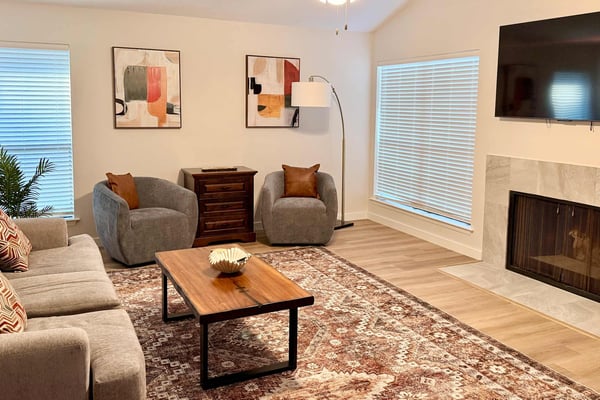 Bright living room with gray seating, wood coffee table, fireplace, wall-mounted TV, and multiple windows with shutters