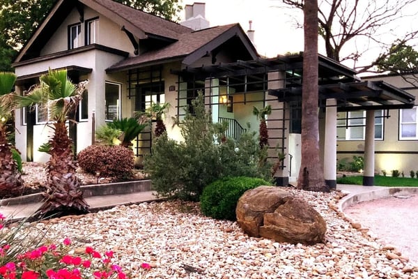 Mid-century modern house with cream and dark trim, gravel front yard with boulders, palm trees, and pink flowering shrubs